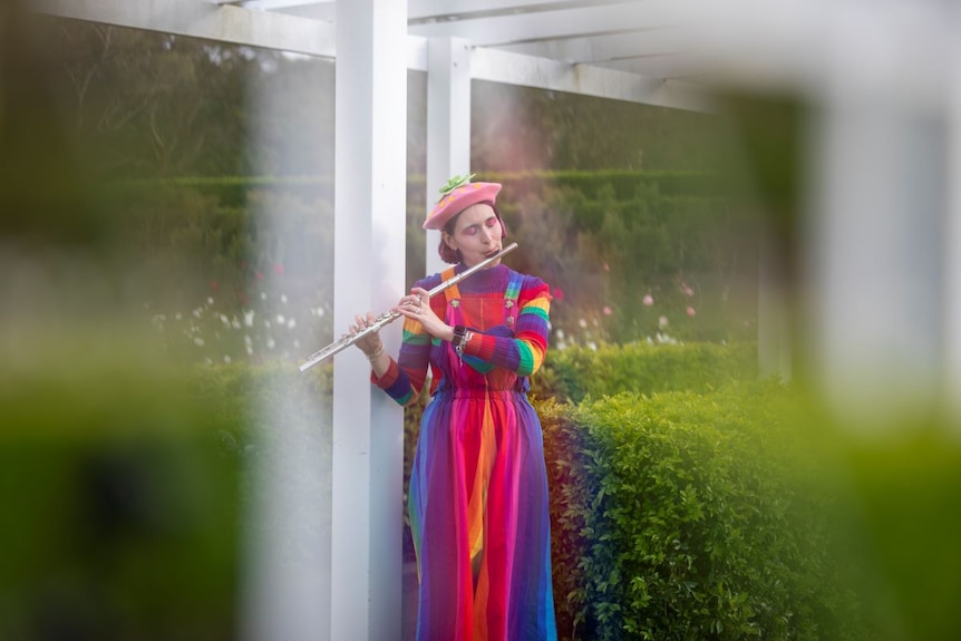 A young woman in pink beret, rainbow jumper and rainbow dress plays a flute in the garden