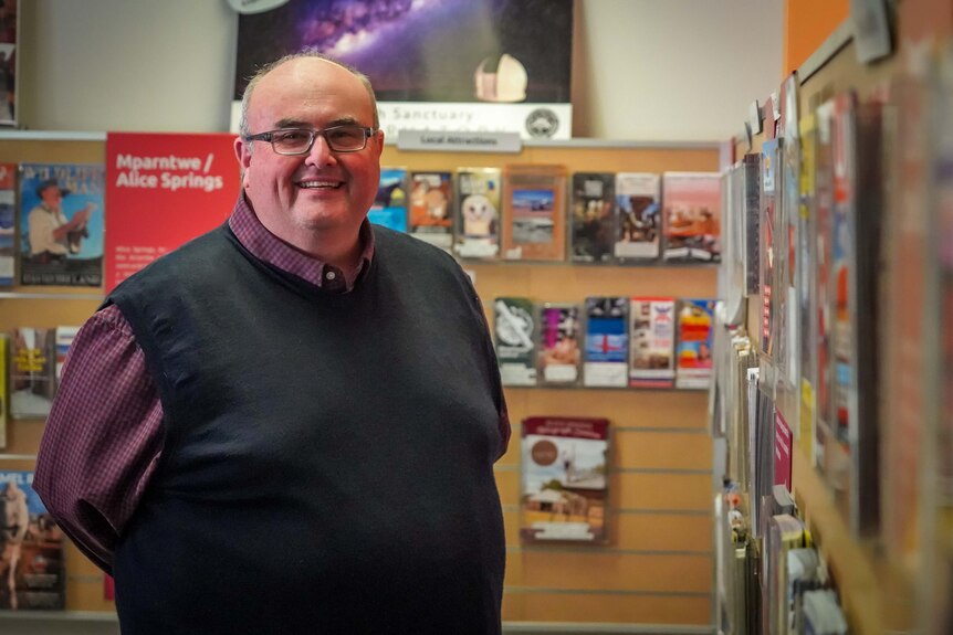A man wearing glasses wears a black shirt in a tourism centre. He's smiling. 