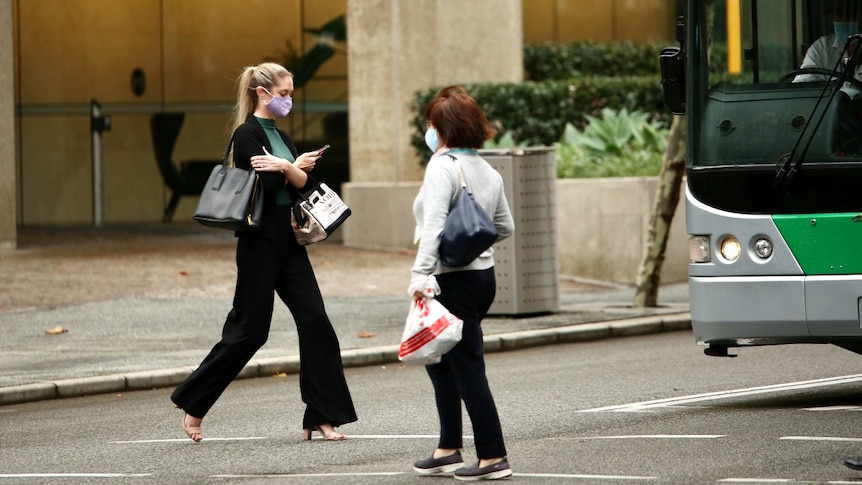 Two women wearing masks cross a road in the Perth CBD with a bus in background.