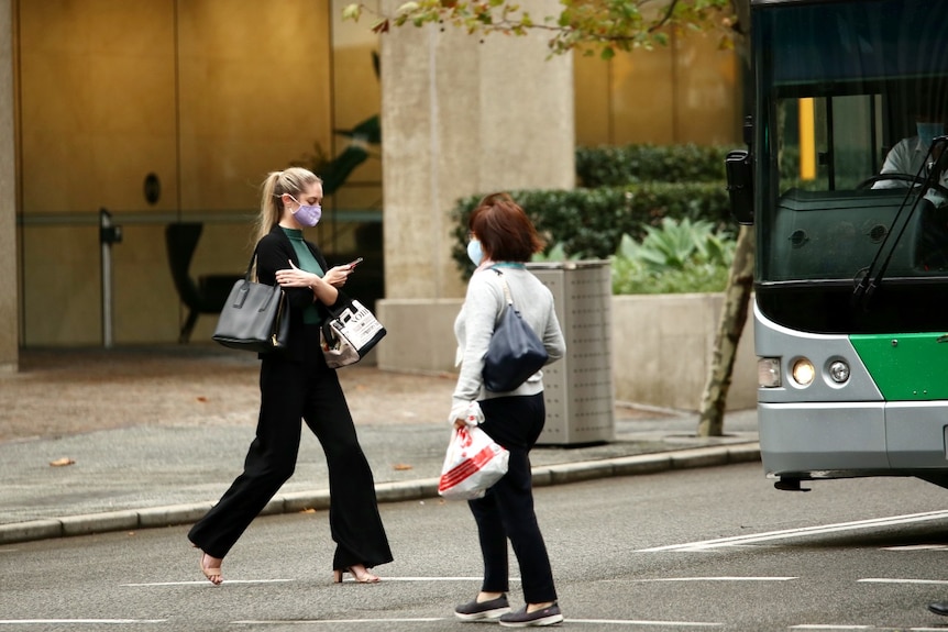 Two women wearing masks cross a road in the Perth CBD with a bus in background.