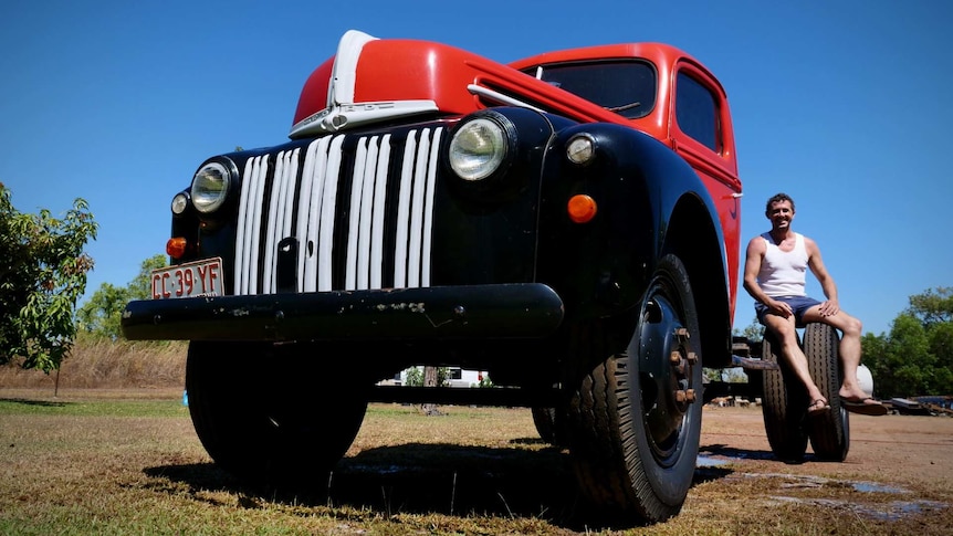 A retro vintage red, black, and white lorry on grass with blue skies with man sitting on the back wheel