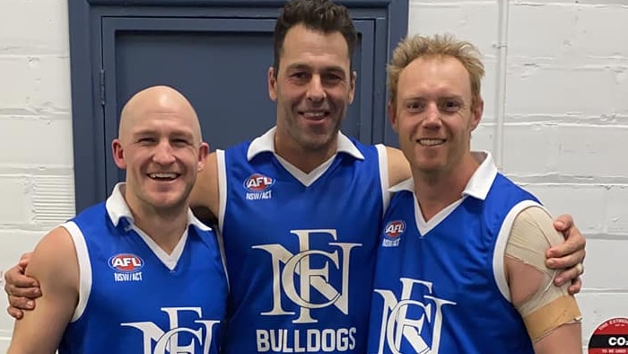 Three middle-aged men wearing blue football jumpers smile for a photo after a match.