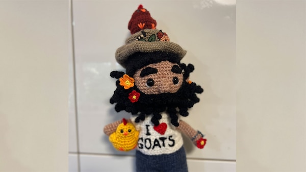 Crocheted doll of Costa holding a yellow chicken and wearing a hat with a brown chicken on top.