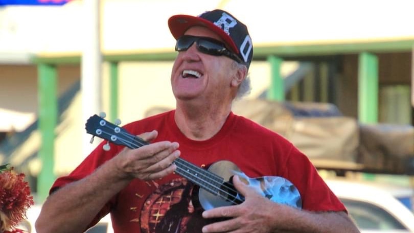 An older man wearing a cap, sunglasses and red t-shirt strums a ukulele.