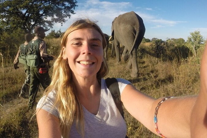 A selfie shot of a young Caucasian woman with blonde hair, outside in the savannah with an elephant and two rangers behind her.