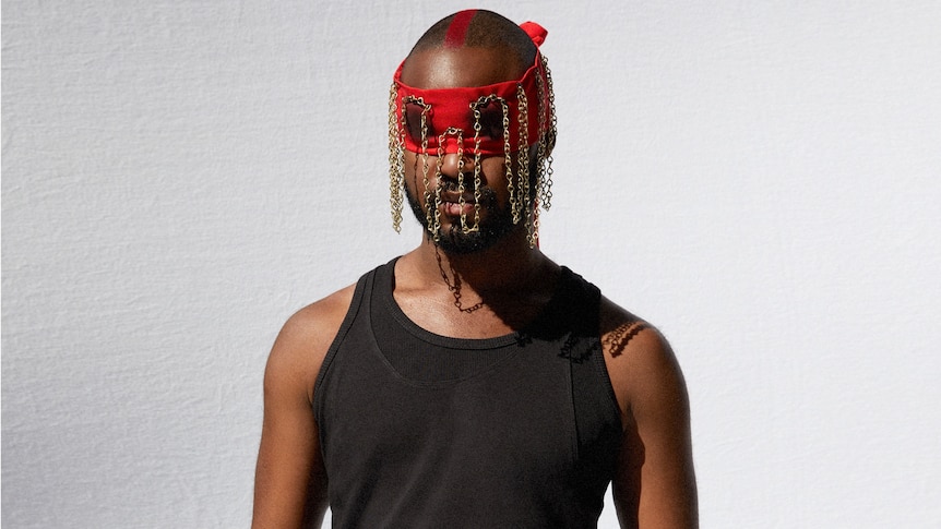 Kofi Owusu-Ansah aka Genesis Owusu stands in a black singlet with a red and gold chained blindfold over his eyes.