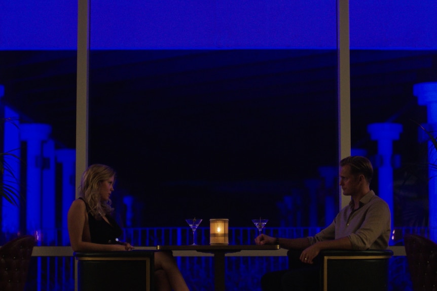 A white woman in a black dress and a white man in a grey shirt sit under intense blue light at a poolside bar drinking cocktails