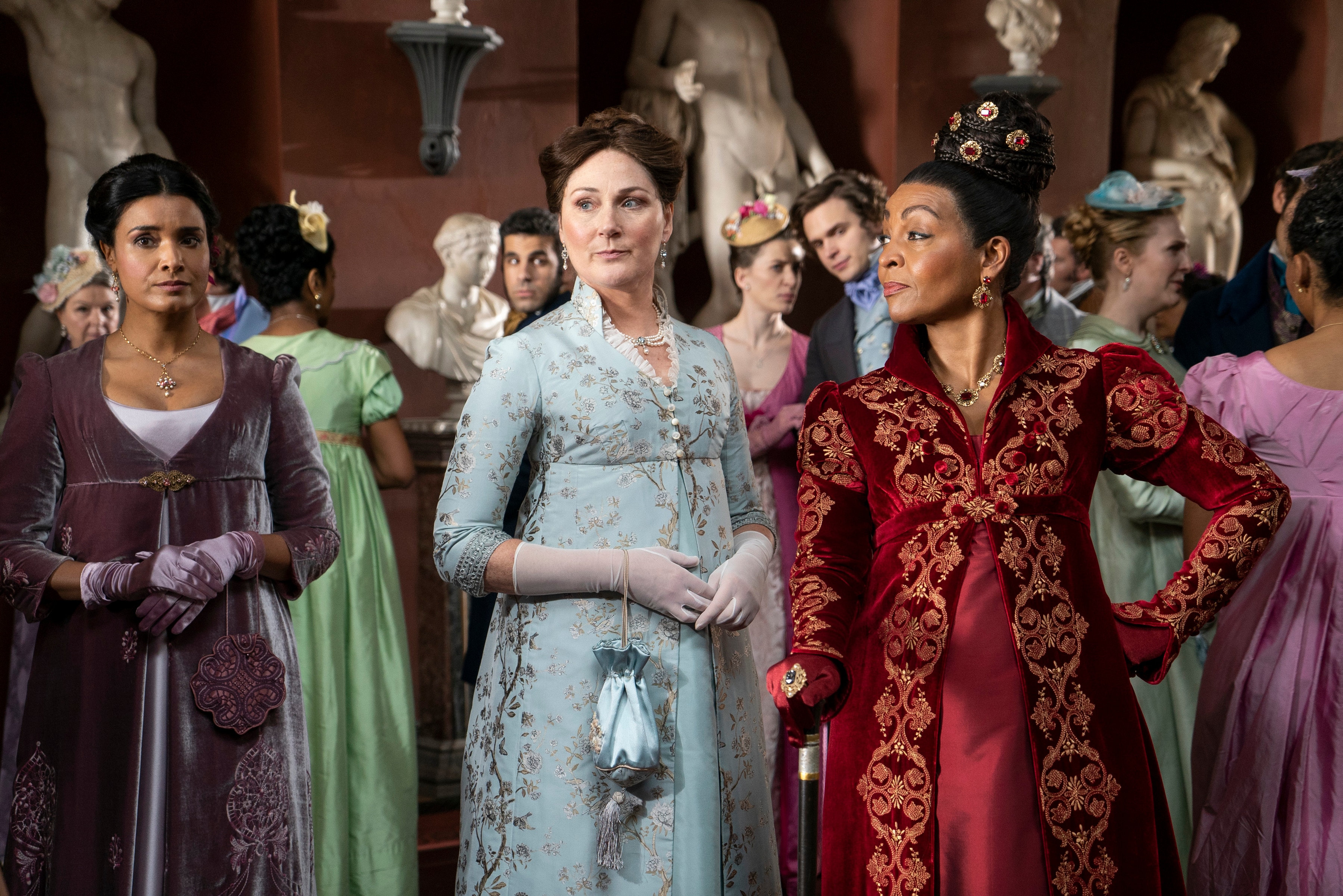 Three women in front of a party of people all wearing colourful period costumes with hair tied back.