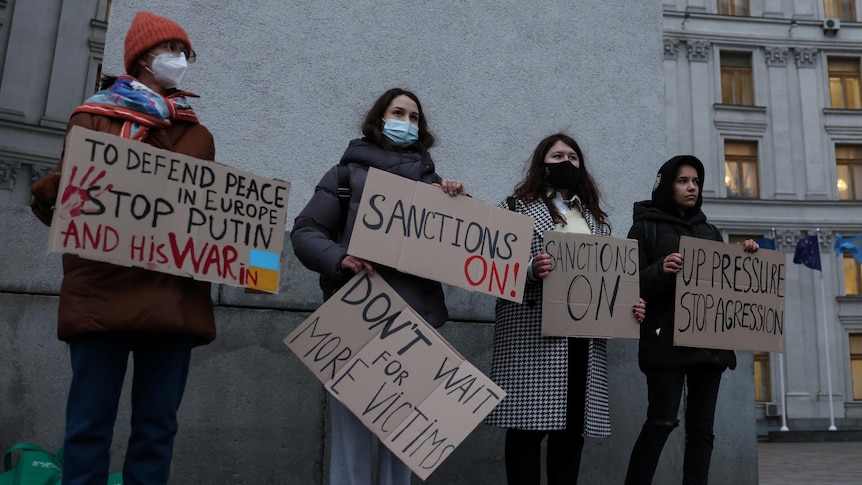 A group of four young people stand outside a grey brutalist-looking building, holding signs for more sanctions on Russia 