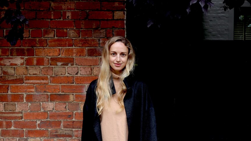 A white woman in her early 30s with blonde hair wearing a black jacket and standing in front of a brick wall