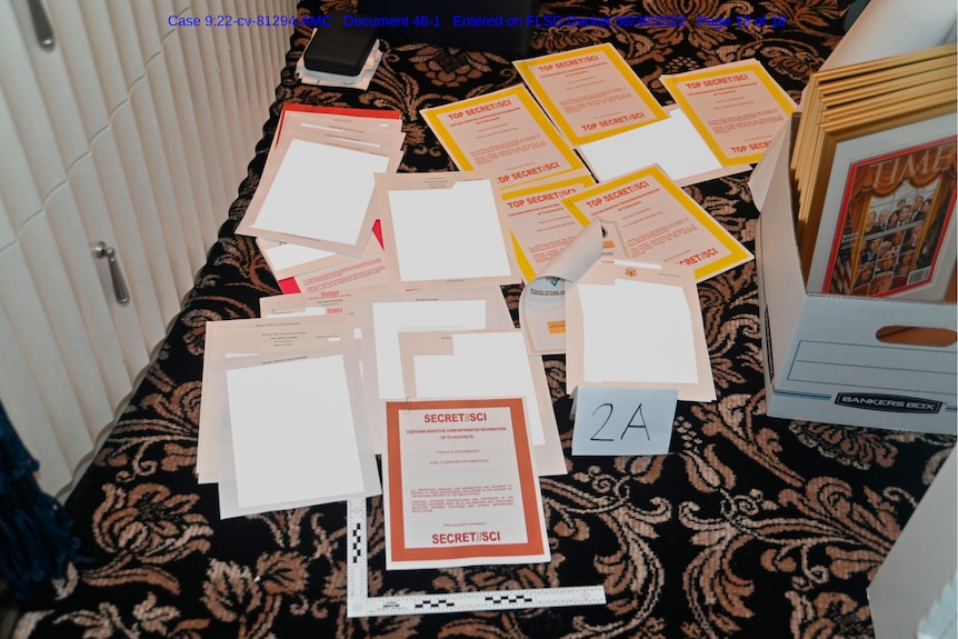 A pile of documents, some redacted to be plain white, others clearly marked Secret and Top Secret, are spread out on carpeting.