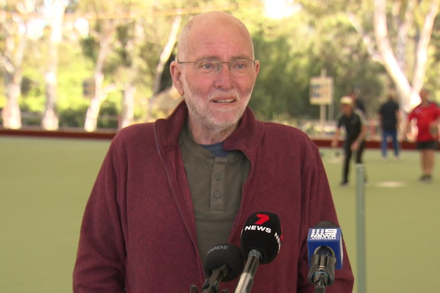 A man with white beard wearing a maroon jumper and khaki top speaking into microphones
