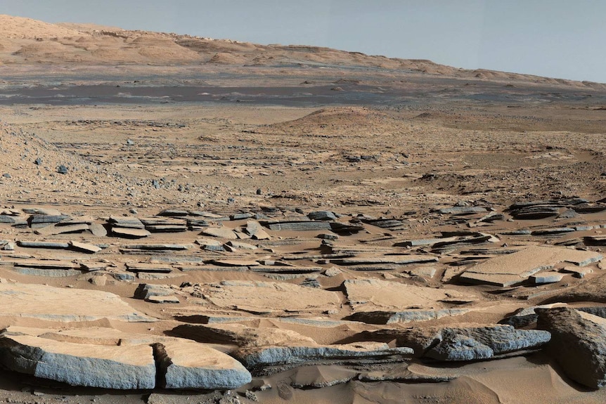 A wide view of the Gale Crater on Mars, showing cracked, flat rocks like a dried-up lake bed