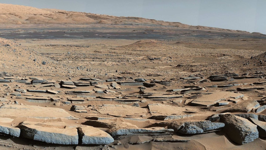 A wide view of the Gale Crater on Mars, showing cracked, flat rocks like a dried-up lake bed