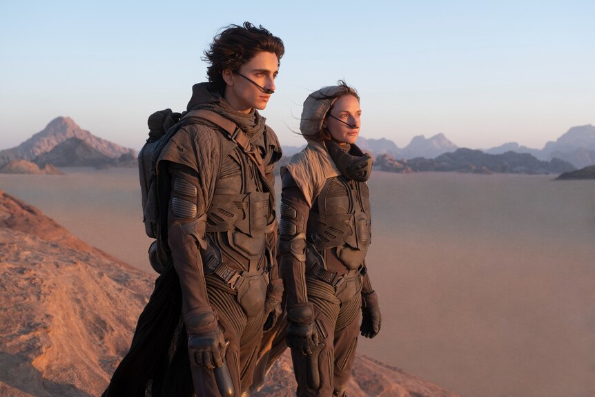 A 20-something man with shaggy dark hair and a woman in her late 30s in a headscarf stand determined in a barren desertscape