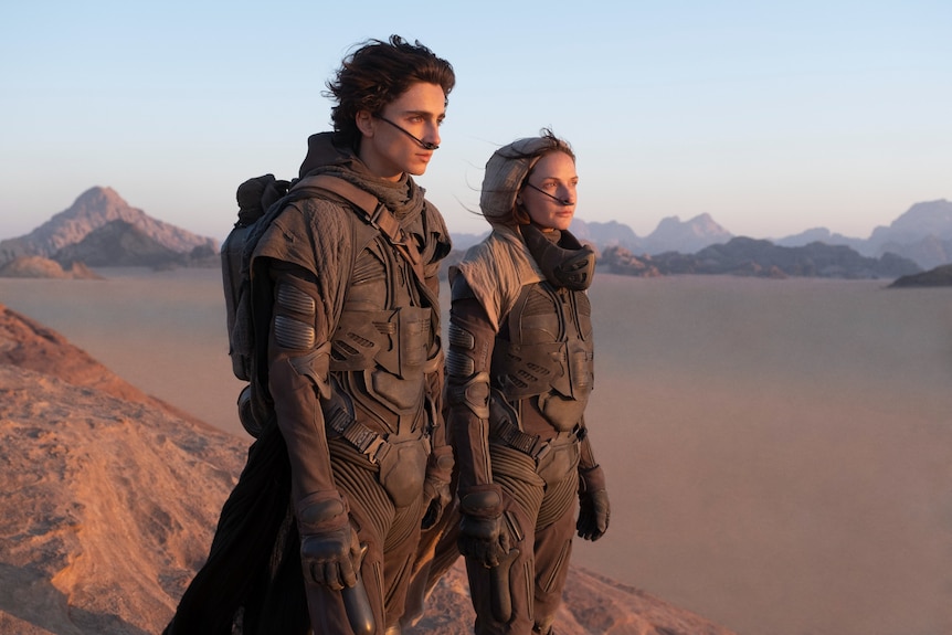 A man in his twenties with unkempt dark hair and a woman in his thirties in a scarf stand determined in a barren desert landscape