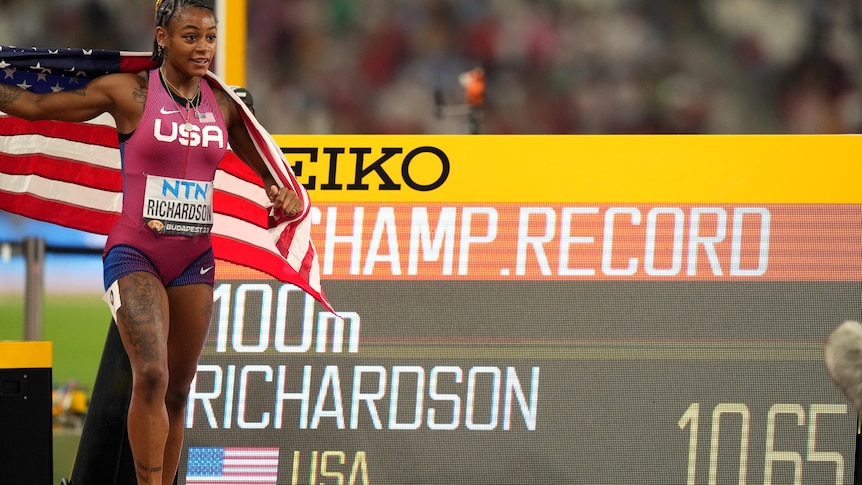 Sha'Carri Richardson, draped in an American flag, stands next to the board with her time and CHAMP RECORD