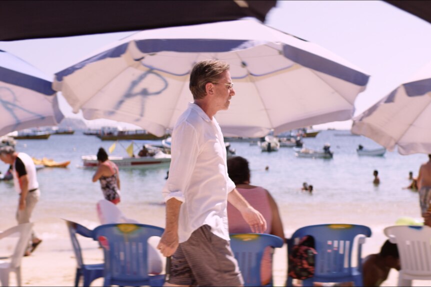 White man with greying beard and short mousey hair wears white shirt and khaki shorts and walks past umbrellas on a beach.