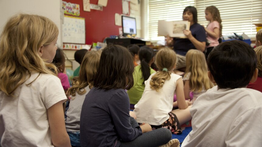 Children wearing casual clothes seen from behind sitting on the floor facing a teacher in the background who holds a book. 