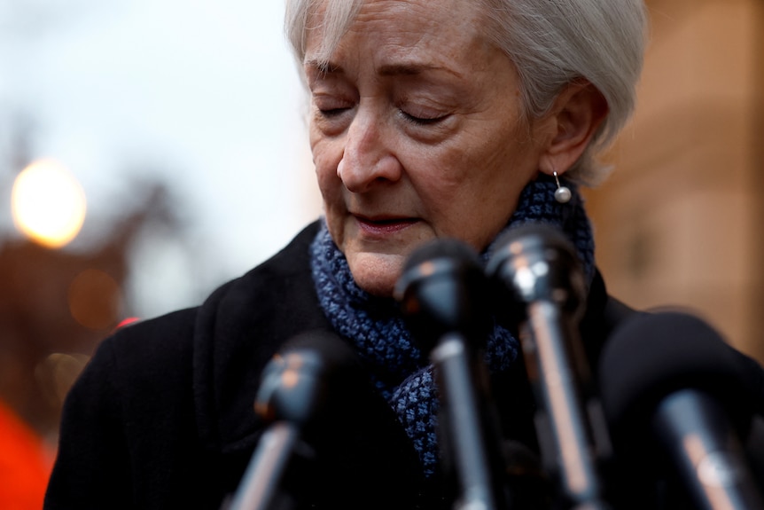 A woman with short grey hair closes her eyes as she speaks into a bank of press mics.