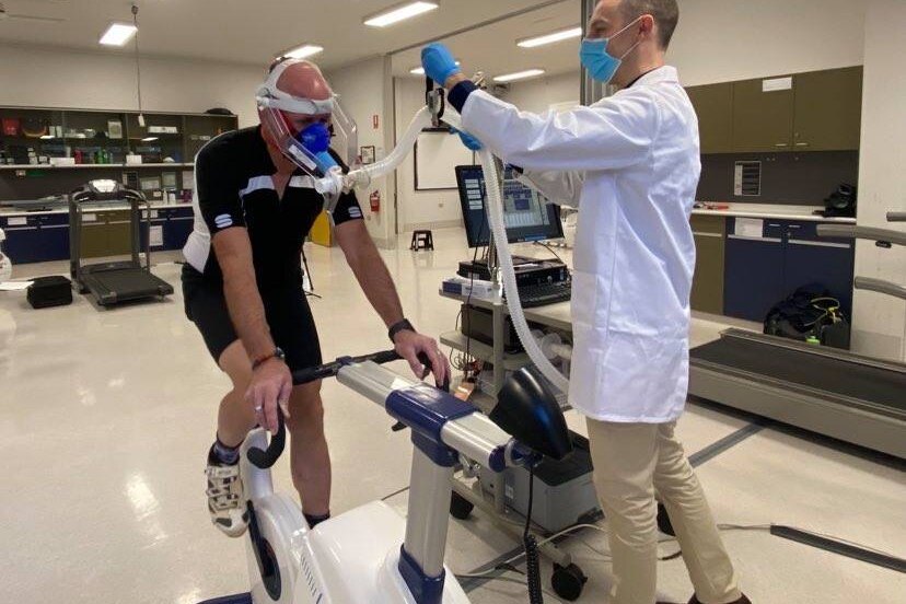 Man in lab coat and face mask sets up equipment while man on bicycle wears oxygen mask