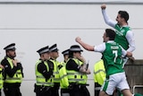 Jamie Maclaren celebrates scoring a goal by punching the air alongside John McGinn as police stand in the background.