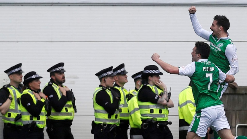 Jamie Maclaren celebrates scoring a goal by punching the air alongside John McGinn as police stand in the background.