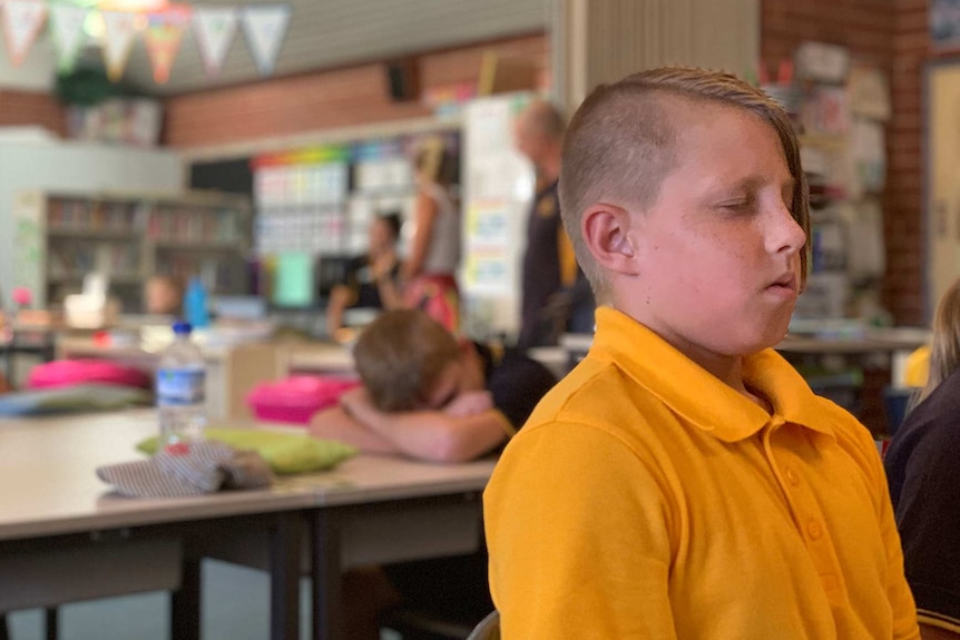 A school boy in a yellow shirt closes his eyes and meditates while sitting at a desk in a classroom.