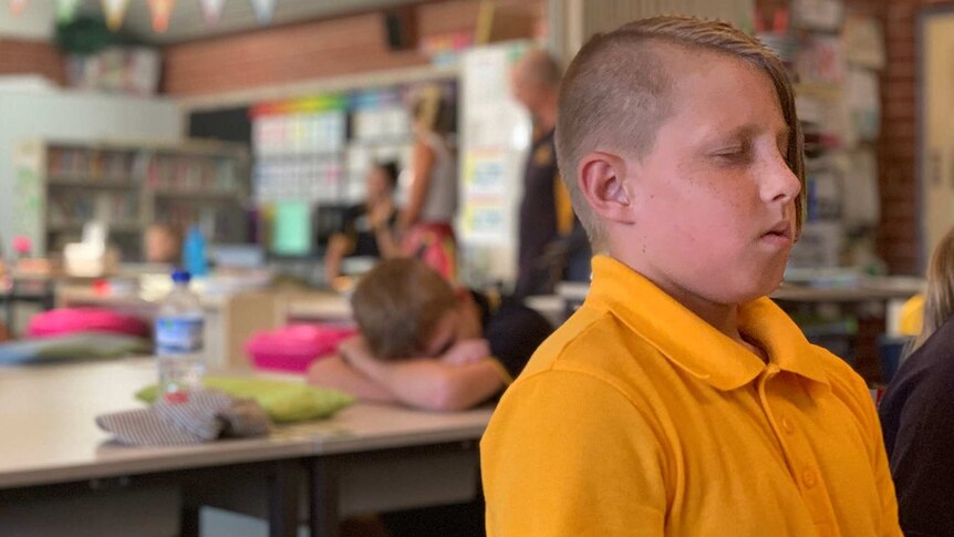 A school boy in a yellow shirt closes his eyes and meditates while sitting at a desk in a classroom.