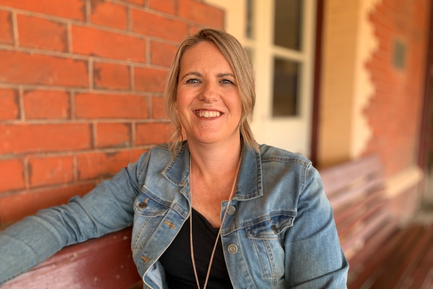 Woman with blonde hair wearing black top and denim jacket sitting on a bench.