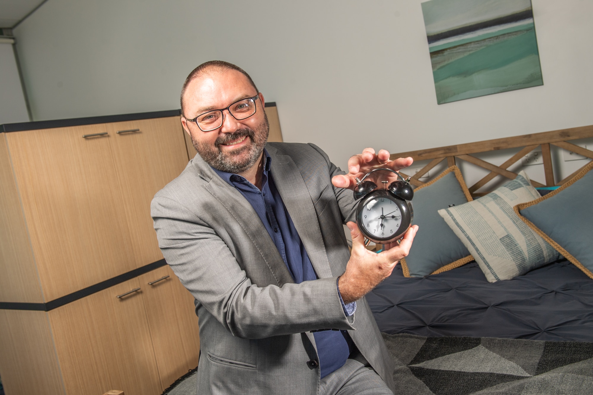 Un hombre con traje se sienta en una cama y sostiene un antiguo despertador
