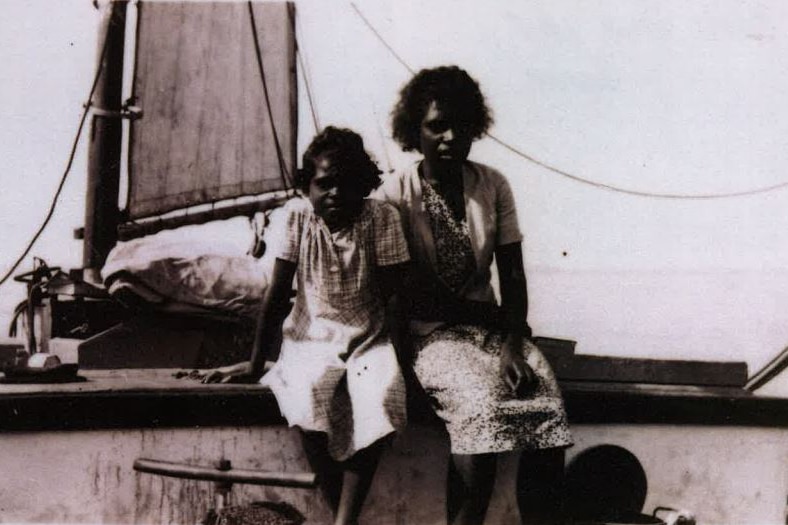 An old black and white image of a young girl sitting on a boat with an older woman perched next to her.