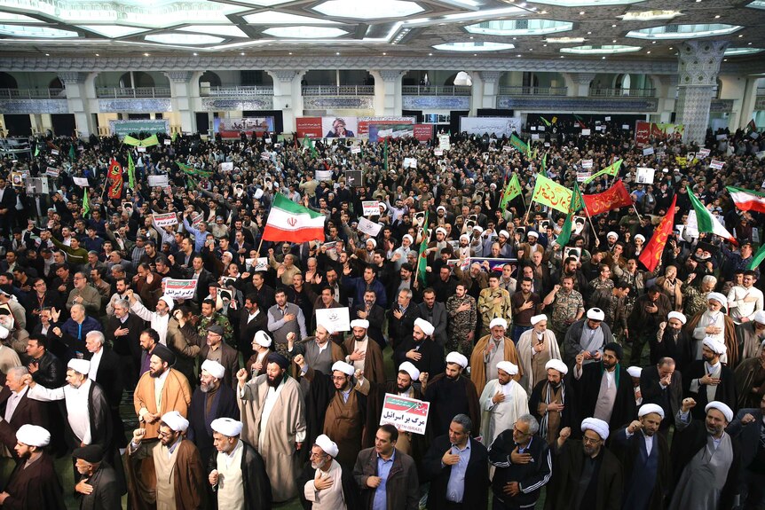 A wide shot of a crowd shows demonstrators waving Iranian flags and chanting with their hands over their hearts.