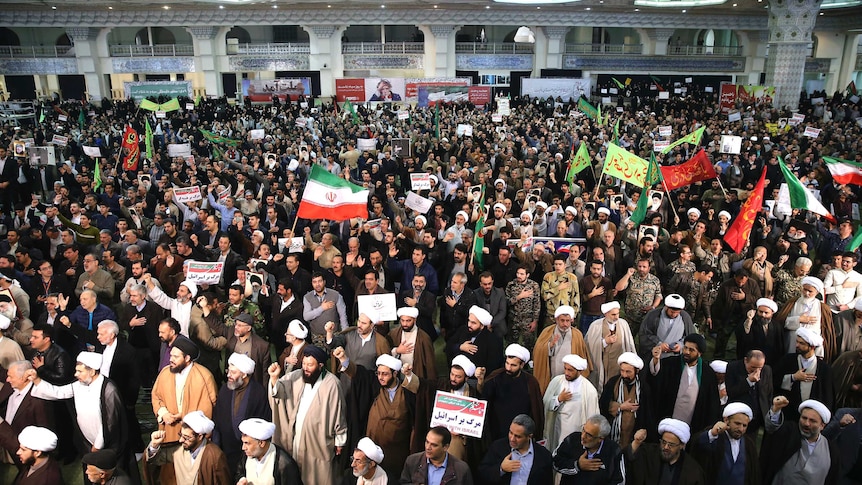 A wide shot of a crowd shows demonstrators waving Iranian flags and chanting with their hands over their hearts.