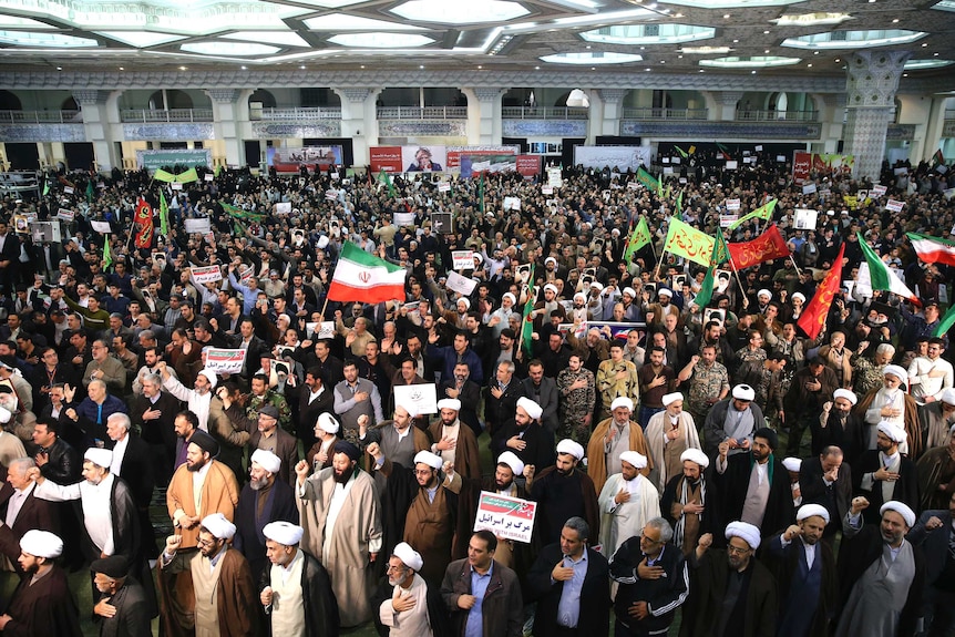 A wide shot of a crowd shows demonstrators waving Iranian flags and chanting with their hands over their hearts.