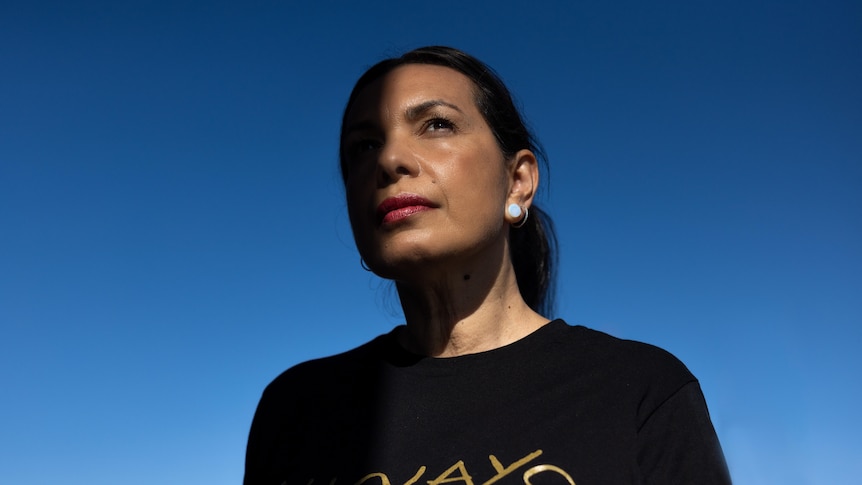 A Kokatha woman with black hair pulled back wears a black shirt that reads 'Always' in gold and looks up against a blue sky.