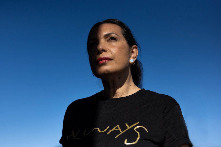 A Kokatha woman with black hair pulled back wears a black shirt that reads 'Always' in gold and looks up against a blue sky.