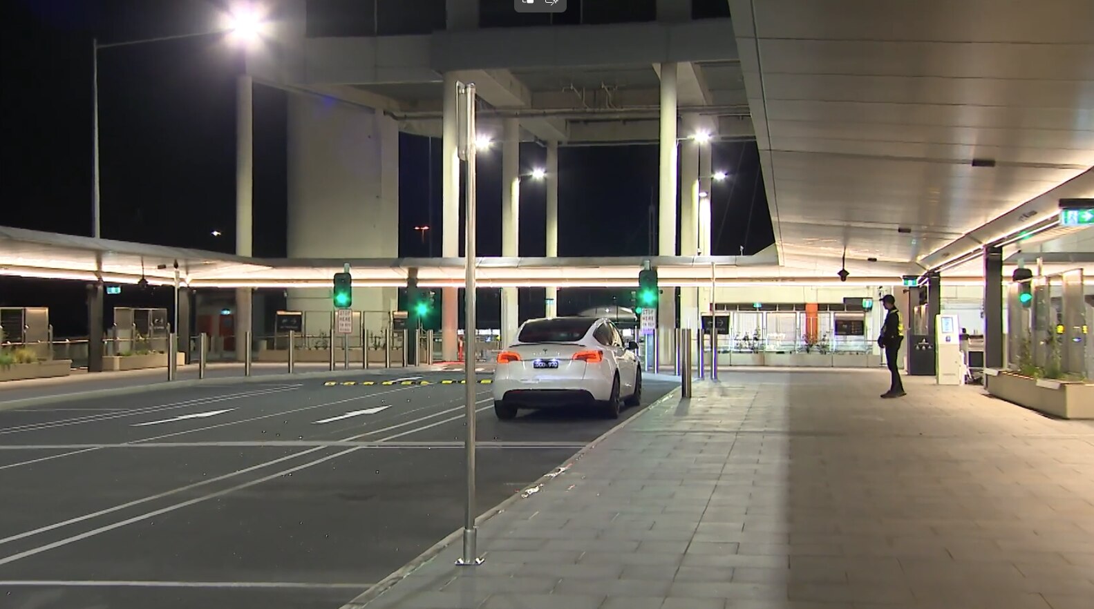 A security guard stands on a footpath beside a white car parked in a bay at night.