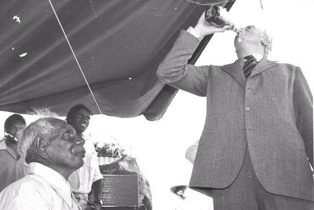 An older white man chugging champagne out of the bottle as an Aboriginal man looks on.
