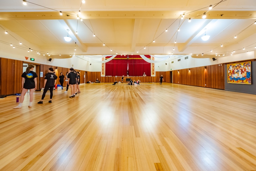 Dancers wait for their practice in a big dance hall with shiny floor boards and lights hanging from the ceiling. 