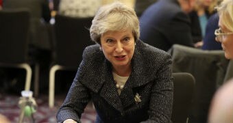 A woman in a dark suit coat places her hands on a table as she leans in to talk to another woman seated at the table.