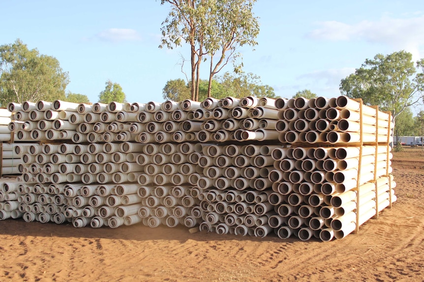 a stack of white piepline on red dirt with trees behind.