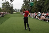 An image taken behind a male golfer as he tees off on the 18th hole at the Masters while watched by a gallery of spectators.