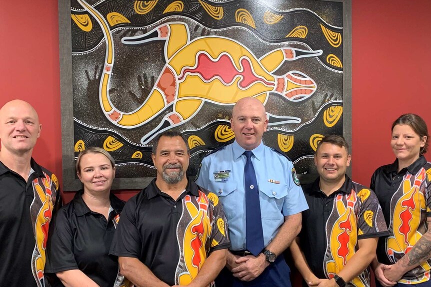 A police officer in uniform stands in front of a Indigenous artwork, flanked by adults wearing shirts also bearing the artwork.