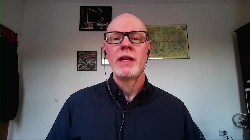 A screenshot from a video conference call shows a man with no hair and black-rimmed glasses.