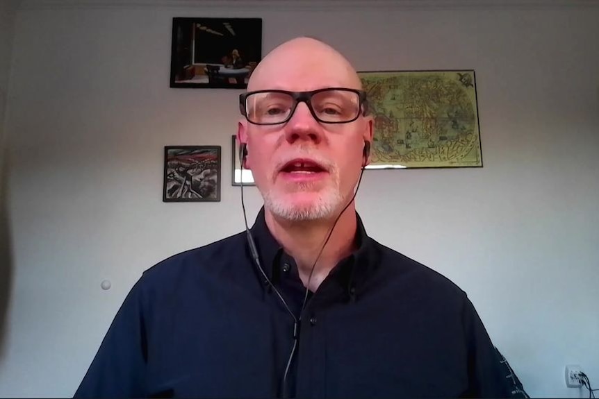 A screenshot from a video conference call shows a man with no hair and black-rimmed glasses.