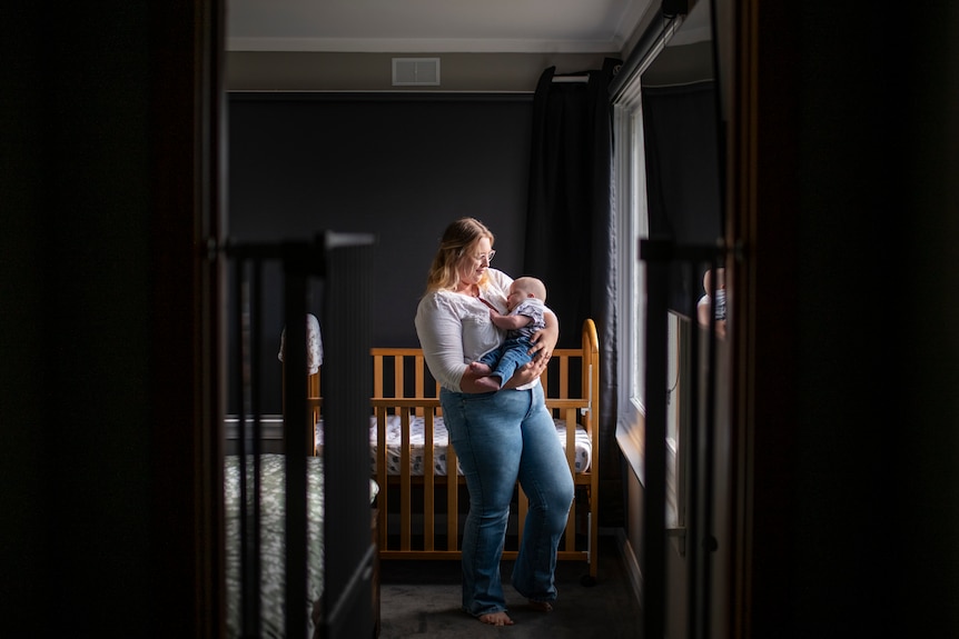 A mother gazes down at her baby in window light, framed in the doorway of her home.