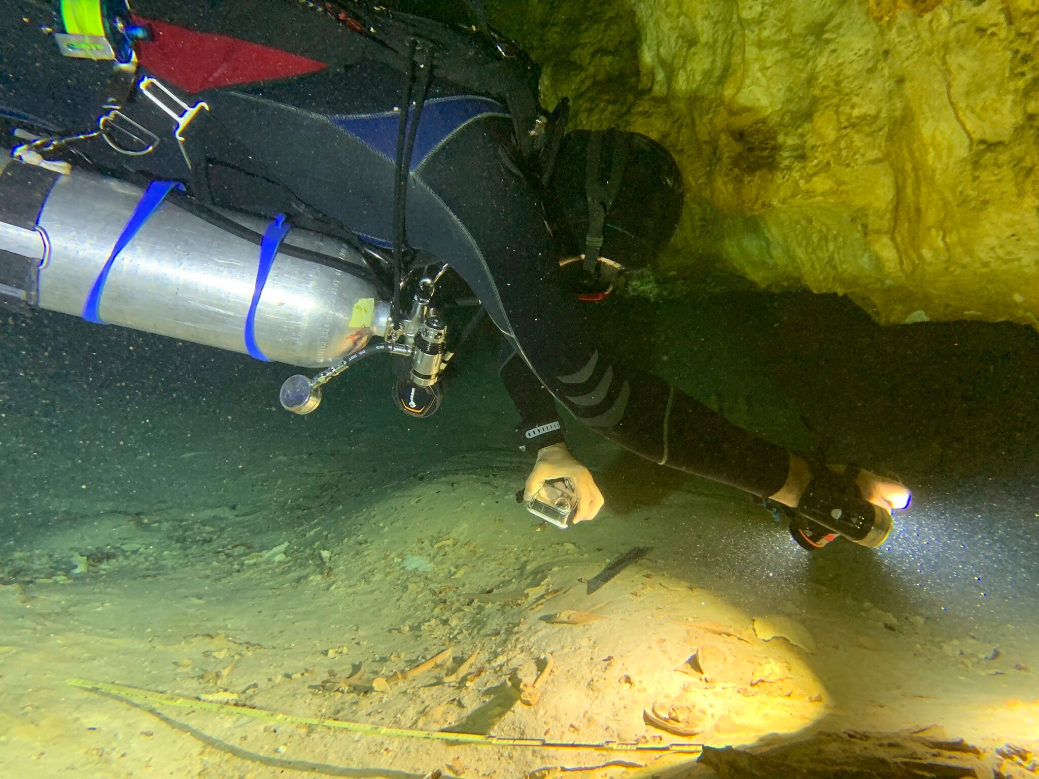 A SCUBA diver uses a torch to illuminate a thin passage between rock and sediment that leads into an underwater cave.
