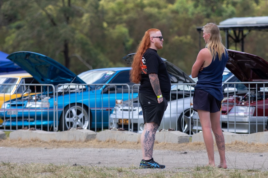 Two men with flowing mullet haircuts stand talking outdoors near some parked cars.