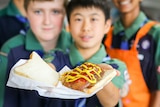 Three young boys in Scouts uniforms hold a sausage sandwich.